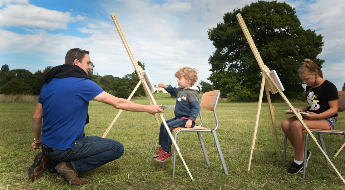 Two children outdoors sat in from on canvases with an adult nearby helping