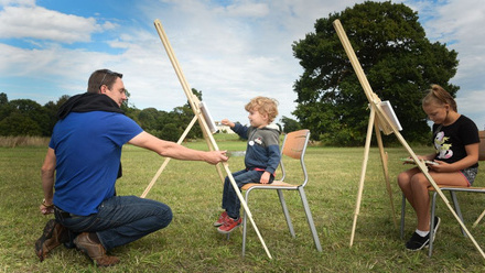 Two children outdoors sat in from on canvases with an adult nearby helping