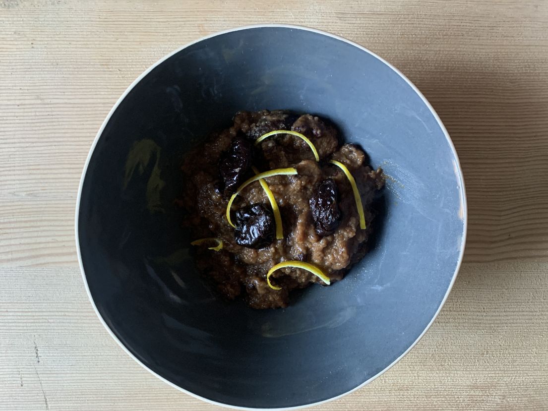 A brown prune and plum pudding in a bowl, garnished by strips of lemon skin.