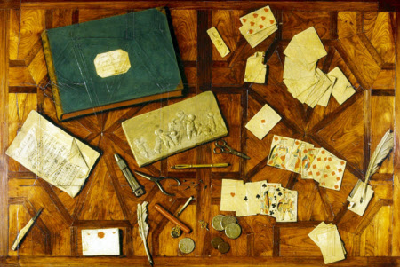 Bird's eye view of a polished wooden table top that looks to be covered with random things: playing cards, letters, coins, quill pen, scissors, book.