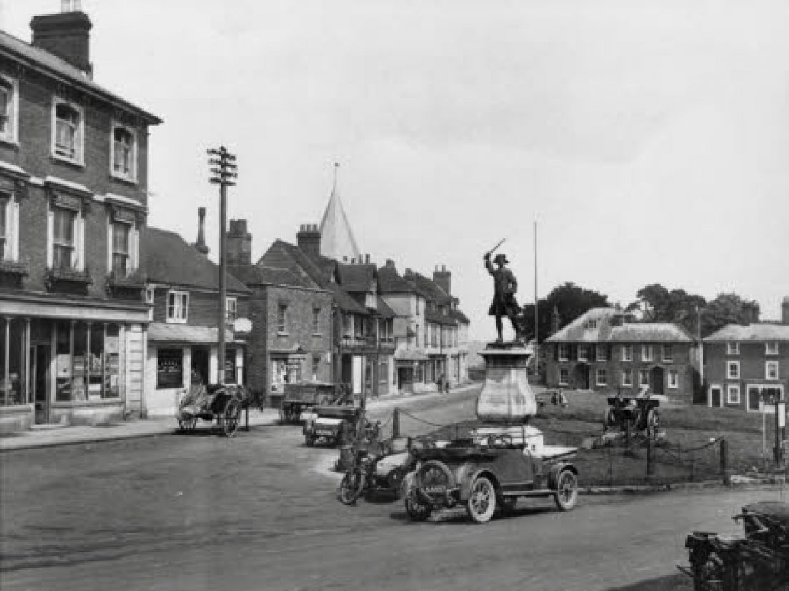 An old black and white photo of a town centre. In a fenced off piece of grass stands a brass statue brandishing a sword.