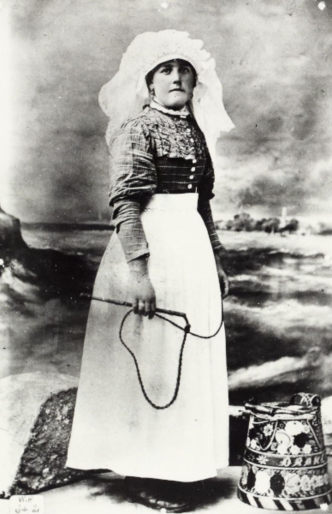 Old photograph of a woman in a balloon sleeved dress with a white apron and a lace headdress. Stood by a painted metal bucket?