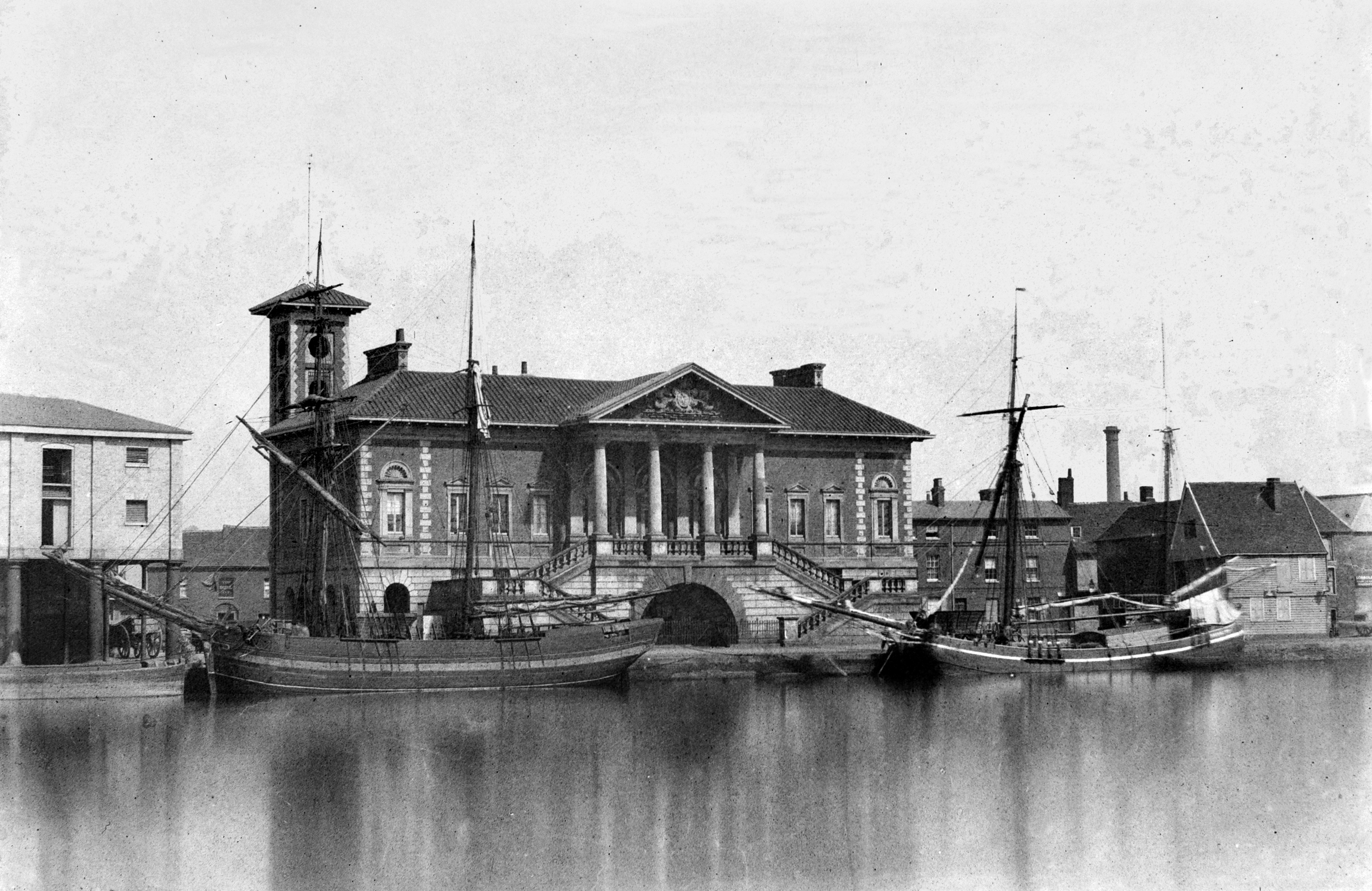 Old photo of a colonnaded building fronting a harbour with two sailing barges docked by it.