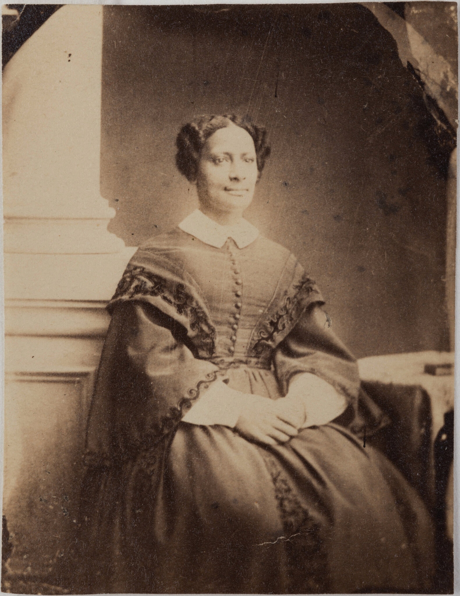 Old photograph of black Victorian woman, seated by a stone pillar, hair up, full skirted dress with embroidered hems and white collar.