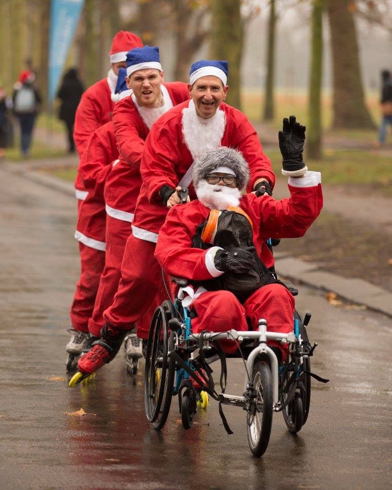4 people in santa suits and in-line skates pushing a 5th person in a santa suit, waving from a wheelchair.