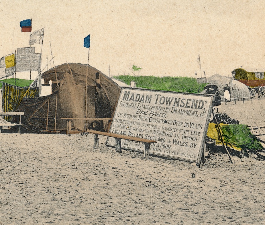 Old, tinted, photograph of camp on a sandy beach. Domed tent with flags outside and a very large sign for Madam Townsend.
