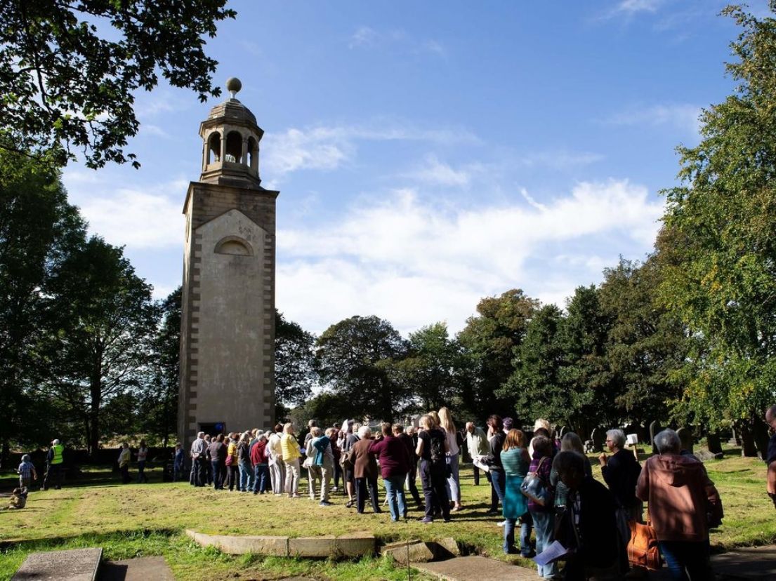 A long queue of visitors lining up through a graveyard towards a stone tower with a bell on top. Surrounding the graveyard are trees.