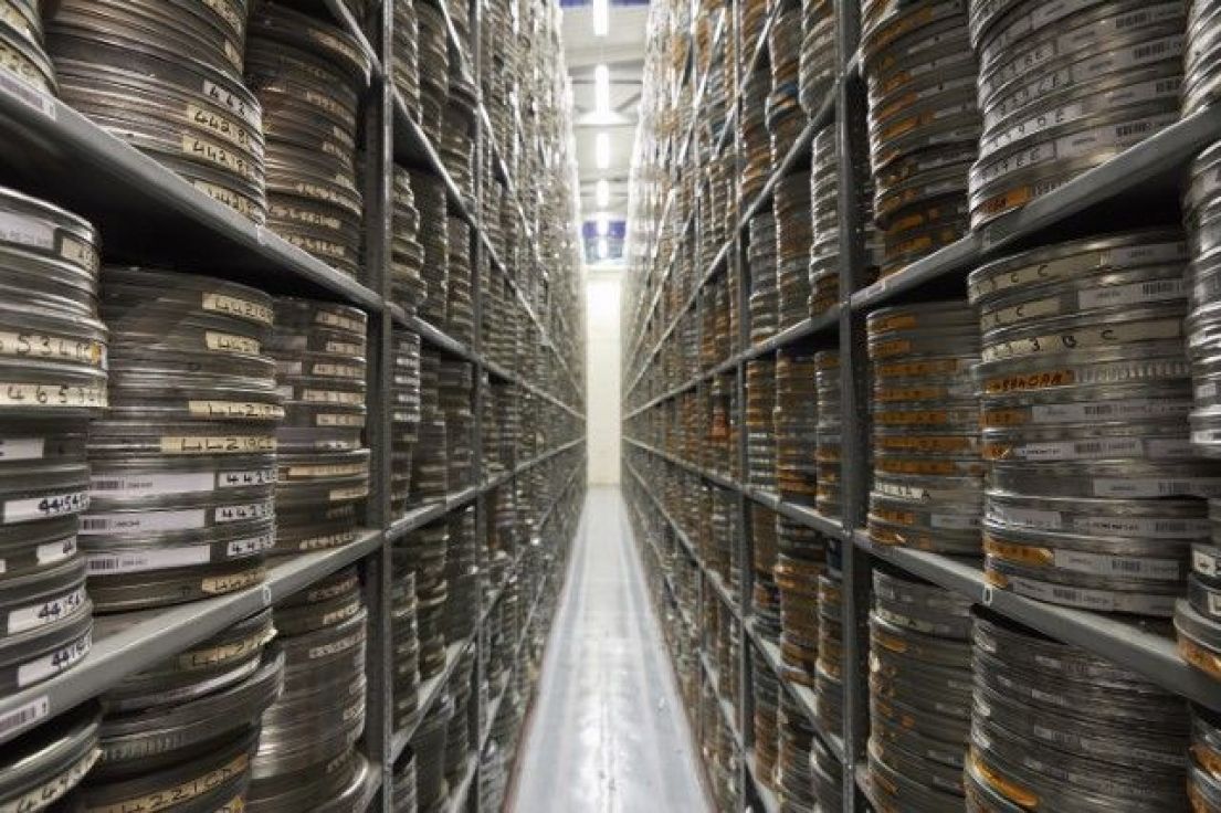 Narrow corridor in a film archive with high shelving stretching away on either side stacked with film canisters
